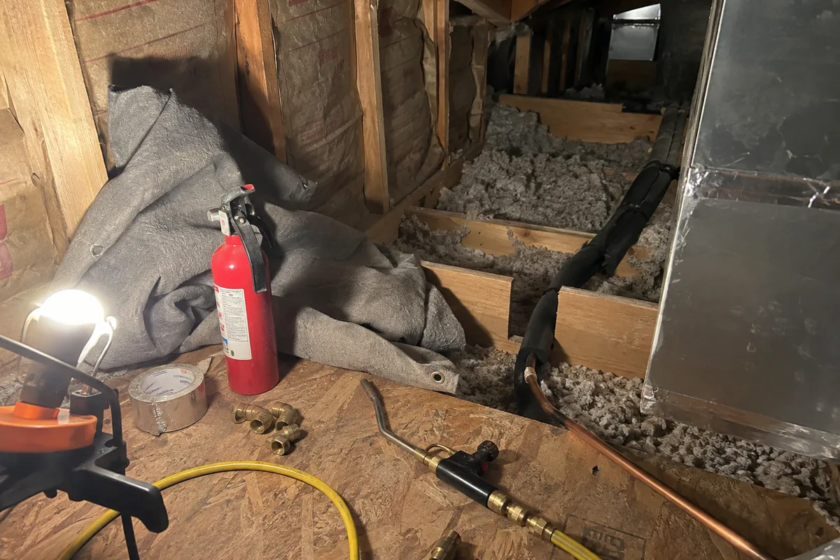 A cramped knee wall attic with kraft faced batt insulation between dry framing, a copper refrigerant line set running through a joist bay, a brazing torch and hose lying on plywood decking beside a small fire extinguisher, a welding blanket bunched against the studs, and a work light throwing hard shadows across blown insulation in the bays.
