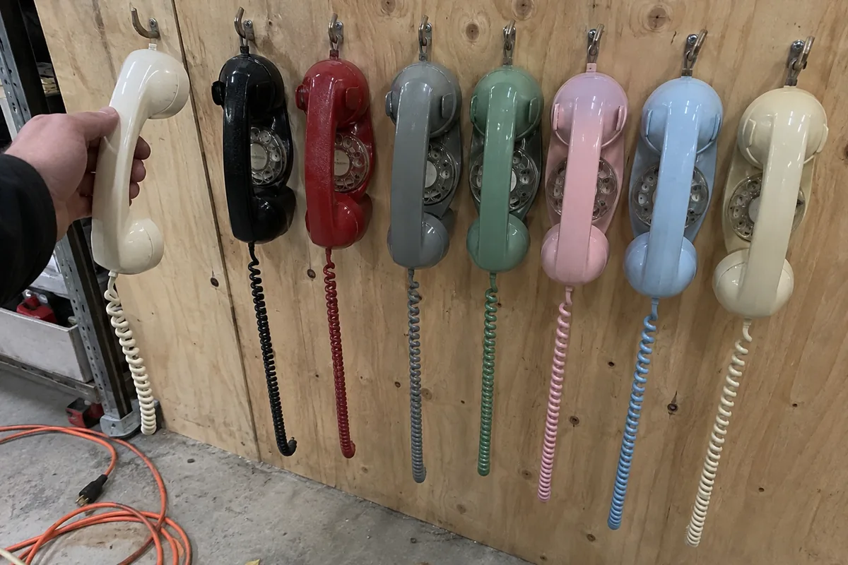 A wall-mounted array of seven vintage rotary phone handsets on hooks against a plain workshop wall, flat overhead light, one handset lifted slightly off its cradle.