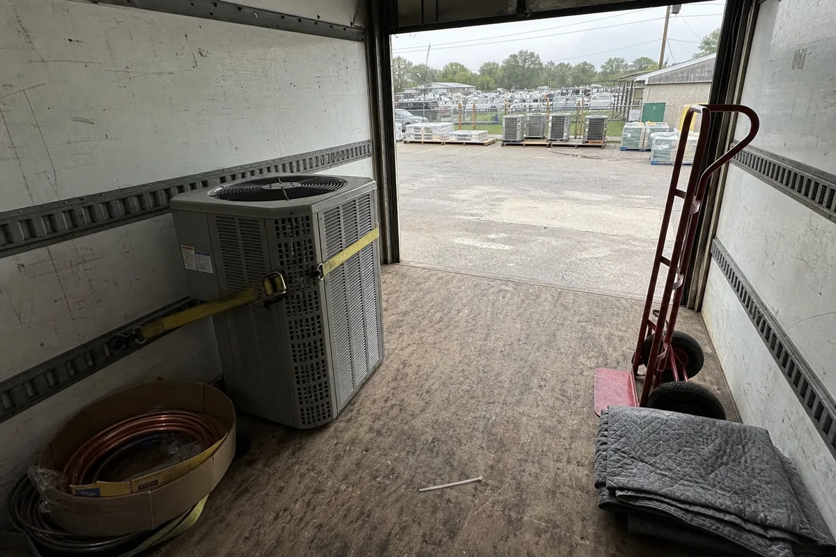 A residential condensing unit strapped upright against the wall of an enclosed box truck, next to a lineset coil, a hand truck, and a stack of moving blankets, with the roll-up door open to a supply house lot.