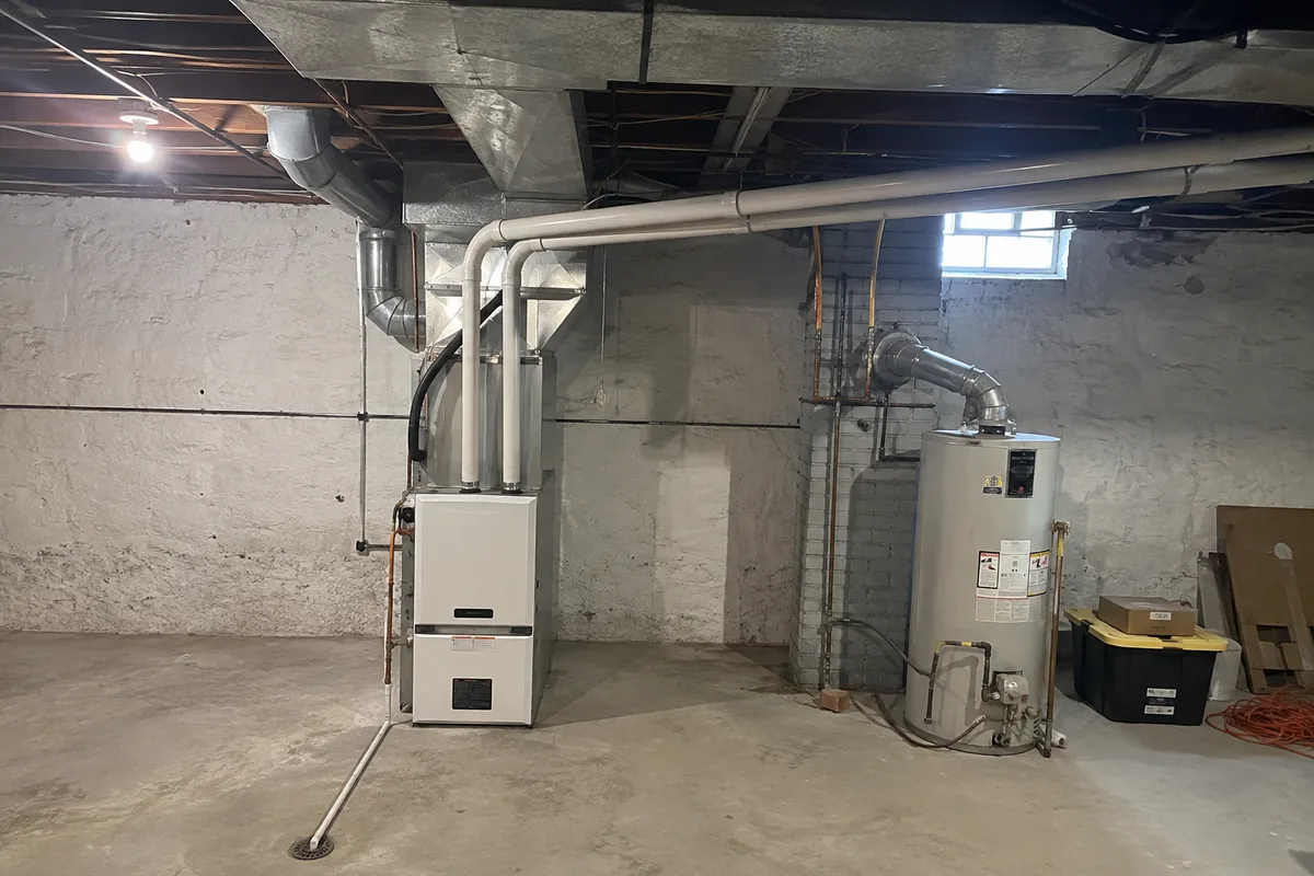 A white condensing gas furnace in a basement with PVC intake and exhaust pipes running through the rim joist, a condensate line to the floor, and an older gas water heater on a metal flue beside it.