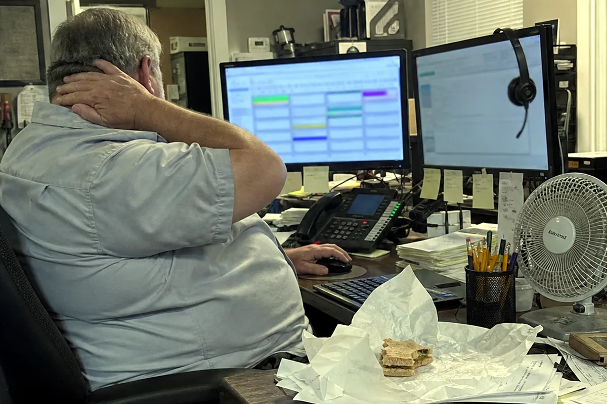 A commercial HVAC dispatcher at a cluttered desk with two monitors, rubbing his neck, a headset and stacked work orders nearby.