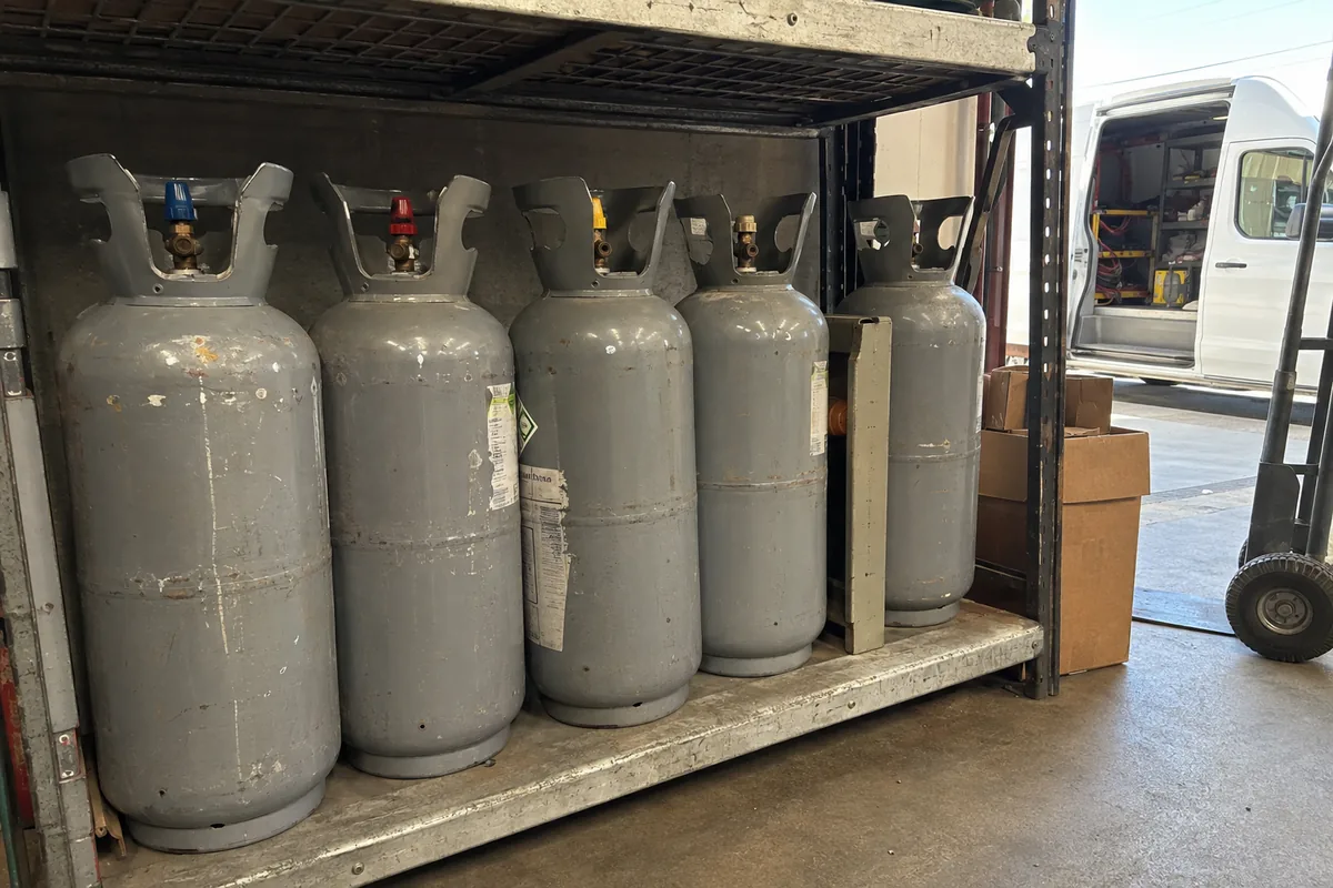 A row of refrigerant cylinders on a warehouse shelf, valve caps visible, with a service van in the background.