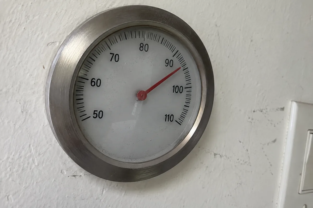 A wall-mounted analog thermostat gauge with a red needle pinned past the 90 degree mark, close crop, flat daylight through a nearby window, dust visible on the glass face.