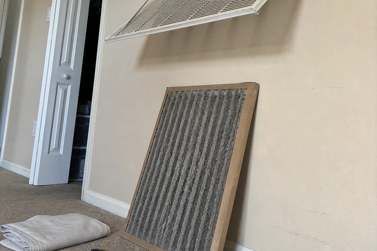 A dust-loaded pleated filter pulled part way out of a hallway return grille, resting against the wall below the open frame.