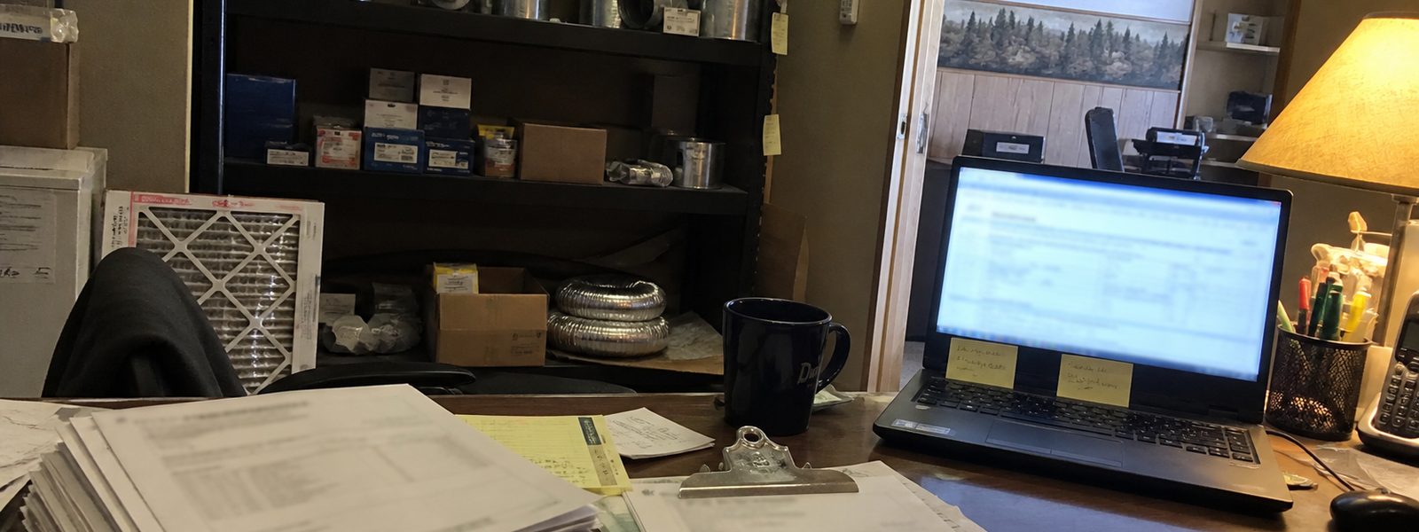 A small HVAC office desk in the evening: laptop, invoices, and a coffee mug under a desk lamp.